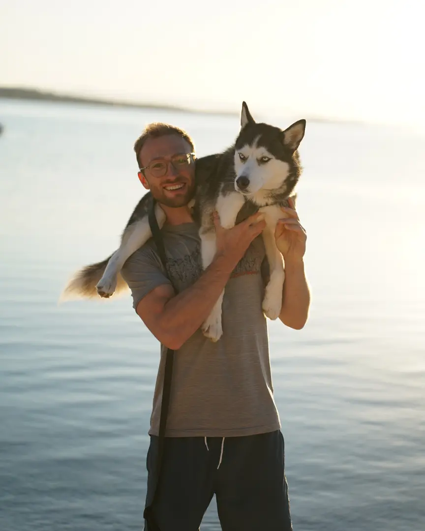 Chris am Wasser im Gegenlicht, hält seinen Husky auf den Schultern